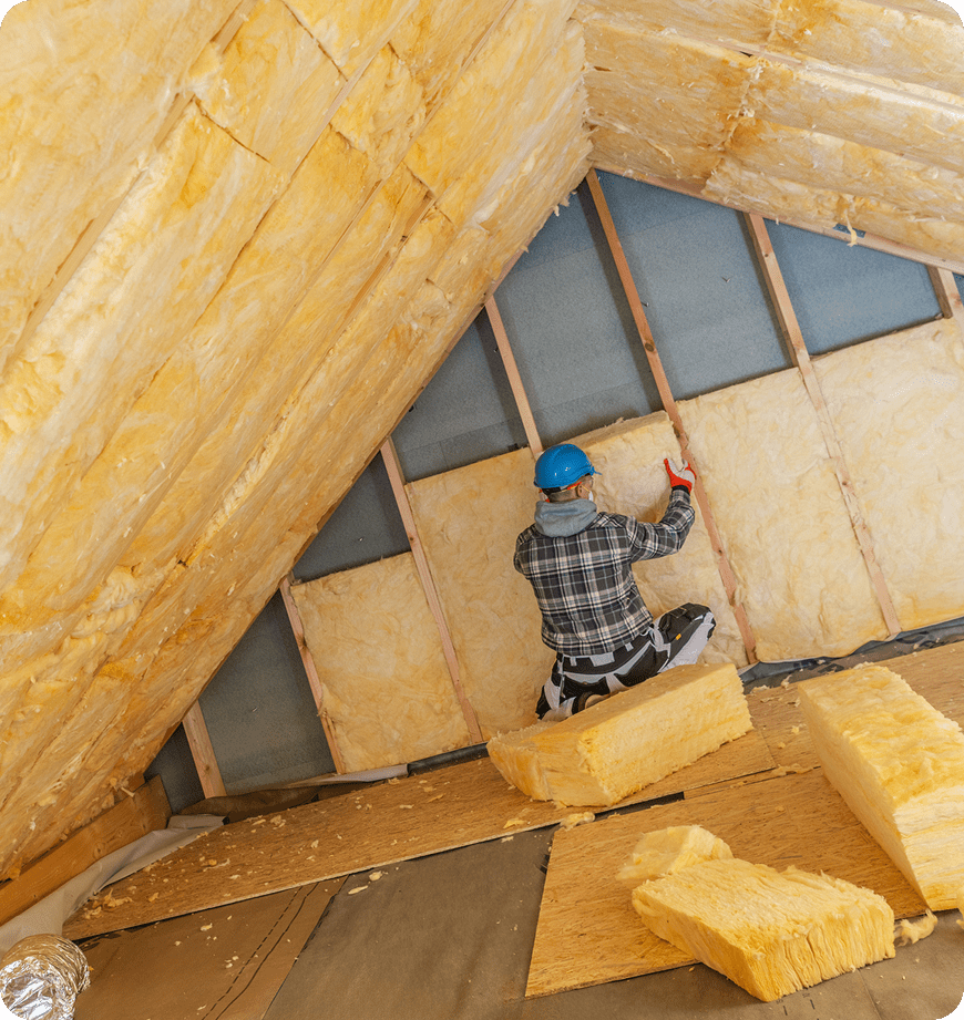 Worker wearing hard hat installing fiberglass insulation in an attic.
